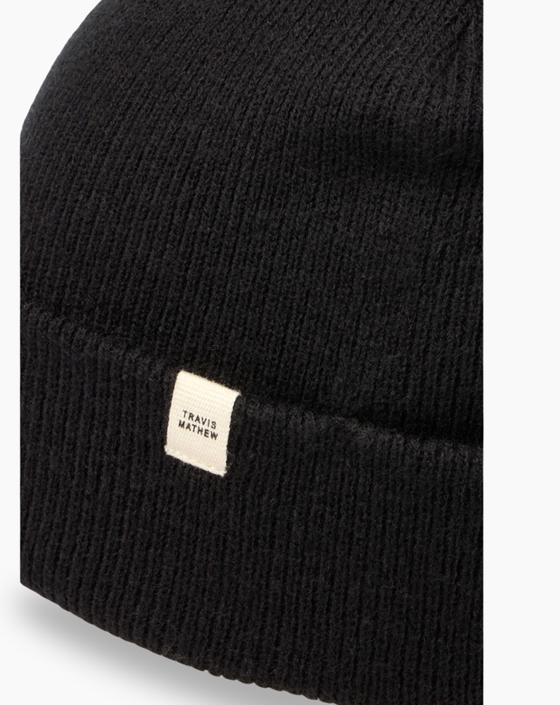 Close-up of the TravisMathew Cloud Beanie in black – rib-knit fabric detail with woven logo label, displayed on a white background.